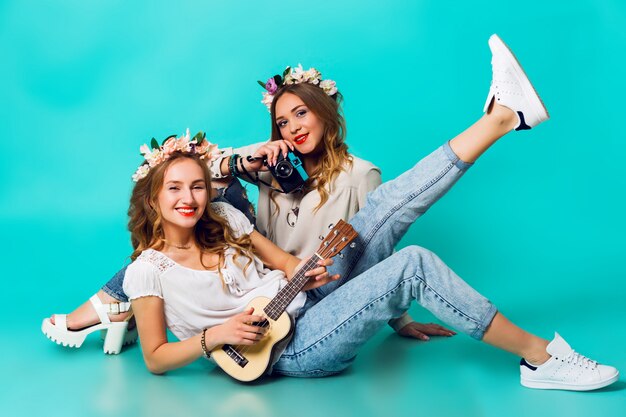 Dos chicas jóvenes divertidas posando sobre fondo de pared azul en traje de estilo de verano con guirnalda de flores con blue jeans y paquete de bolso boho. .