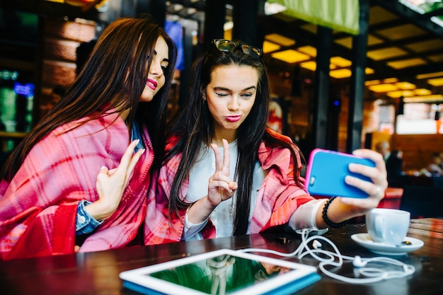 Dos chicas jóvenes y hermosas sentadas en la mesa y haciendo selfie en el café