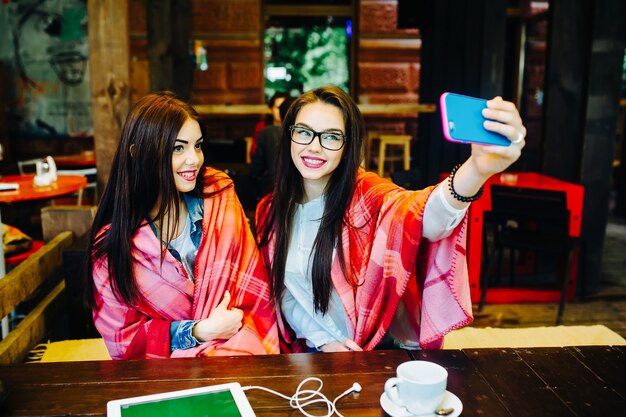 Dos chicas jóvenes y hermosas sentadas en la mesa y haciendo selfie en el café