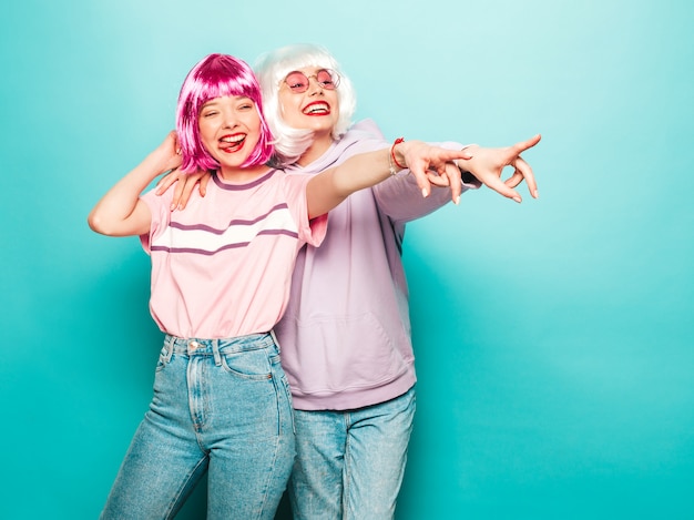 Dos chicas jóvenes hipster sexy con pelucas y labios rojos. Hermosas mujeres de moda en ropa de verano. Modelos despreocupados posando junto a la pared azul en el estudio apuntando a las ventas de la tienda