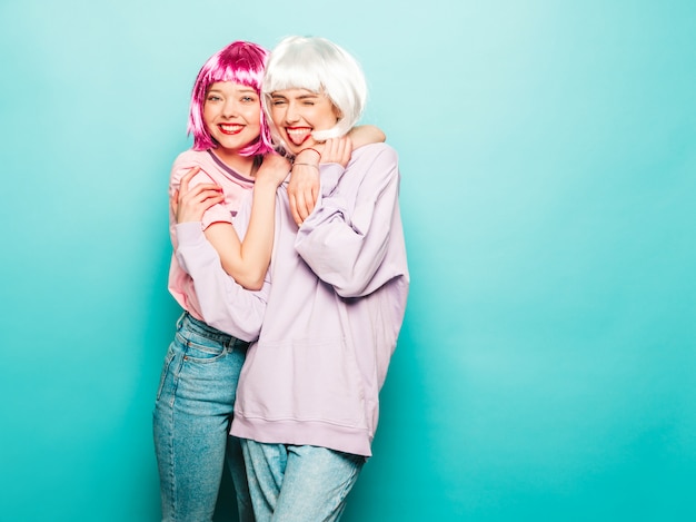 Dos chicas jóvenes y sexy hipster con pelucas y labios rojos. Hermosas mujeres de moda en ropa de verano. Modelos despreocupadas posando junto a la pared azul en el estudio enloqueciendo. Muestra lengua