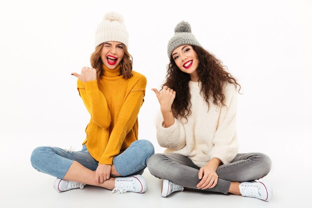 Dos chicas juguetonas con suéteres y sombreros sentados juntos en el suelo mientras apuntan sobre la pared blanca