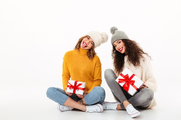 Dos chicas juguetonas en suéteres y sombreros sentados con regalos en el piso juntos sobre la pared blanca