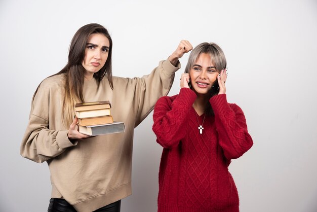 Dos chicas con libro de pie y posando sobre una pared blanca.