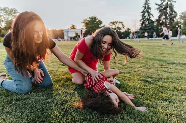 Dos chicas morenas jugando con su hermana pequeña mientras están sentados en el césped. Foto al aire libre de damas pasando tiempo con niños en fin de semana.