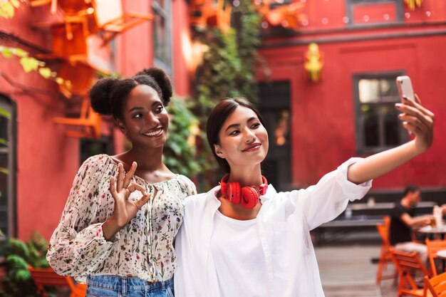 Dos chicas muy elegantes tomando fotos en el teléfono celular mientras pasan felizmente juntas en un acogedor patio