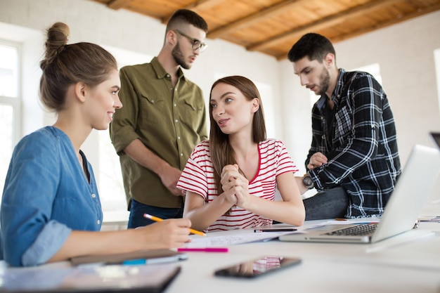 Dos chicas muy sonrientes hablando alegremente mientras pasan tiempo en una oficina moderna con colegas en el fondo Grupo de personas creativas trabajando juntas