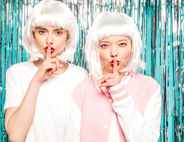 Dos chicas sexy hipster jóvenes con pelucas blancas y labios rojos. hermosas mujeres de moda en ropa de verano. modelos posando sobre fondo azul plata brillante oropel en estudio. muestra el dedo silencio signo de silencio, gesto