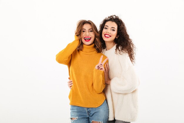 Dos chicas sonrientes en suéteres posando juntos mientras una mujer mostrando un gesto de paz sobre la pared blanca