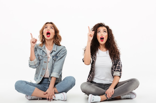 Dos chicas sorprendidas sentadas juntas en el piso mientras señalan y miran con la boca abierta sobre la pared blanca