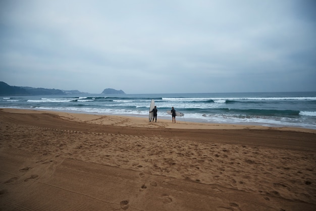 Foto gratuita dos chicas surfistas irreconocibles con sus longboards permanecen en la orilla del océano y observan las olas temprano en la mañana, vistiendo trajes de neopreno completos y listas para surfear