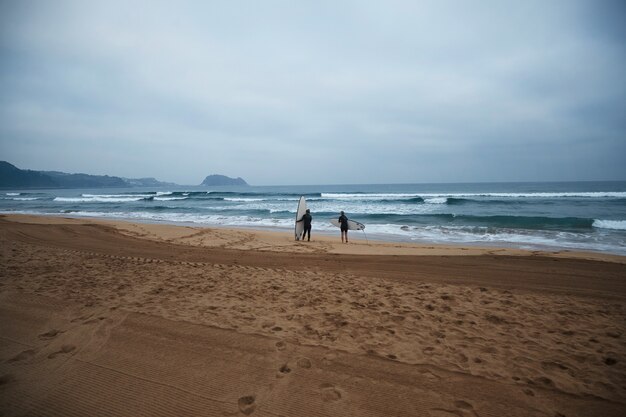 Dos chicas surfistas irreconocibles con sus longboards permanecen en la orilla del océano y observan las olas temprano en la mañana, vistiendo trajes de neopreno completos y listas para surfear