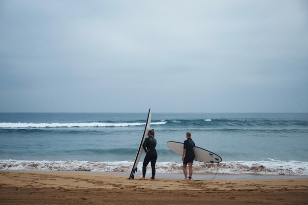 Dos chicas surfistas irreconocibles con sus longboards permanecen en la orilla del océano y observan las olas temprano en la mañana, vistiendo trajes de neopreno completos y listas para surfear