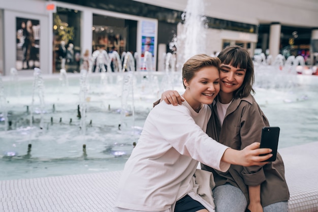 Dos chicas toman una selfie en el centro comercial, una fuente en el fondo