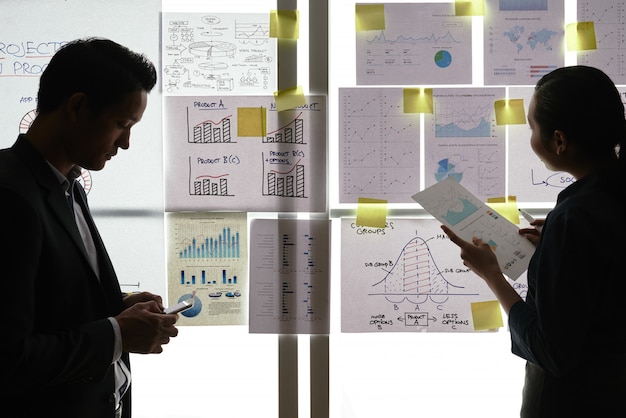 Dos colegas asiáticos de pie junto a la ventana con gráficos y diagramas