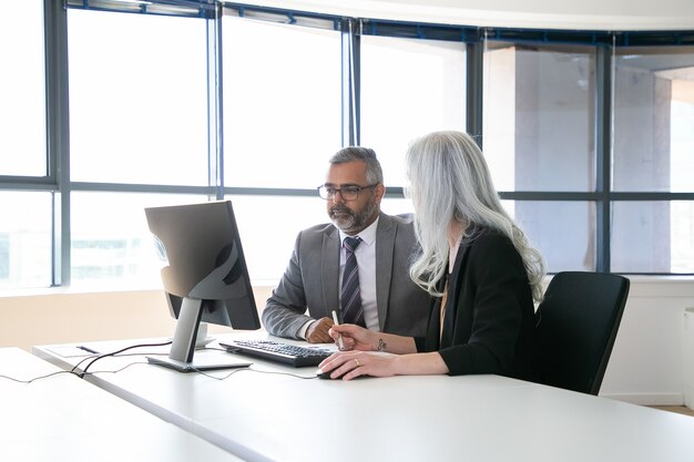 Dos colegas enfocados viendo y discutiendo contenido en el monitor de la computadora, sosteniendo el lápiz y el mouse y hablando mientras están sentados en la sala de reuniones con ventana panorámica. Concepto de comunicación empresarial