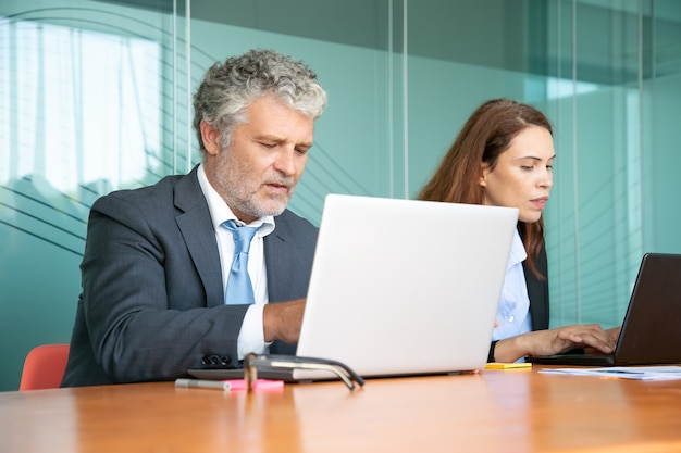 Dos colegas sentados juntos y usando computadoras en la oficina. Empleados de diferentes edades escribiendo en teclados de portátiles.