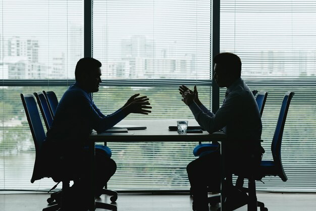 Dos contornos humanos contra la ventana de la oficina cerrada, sentados uno frente al otro y negociando