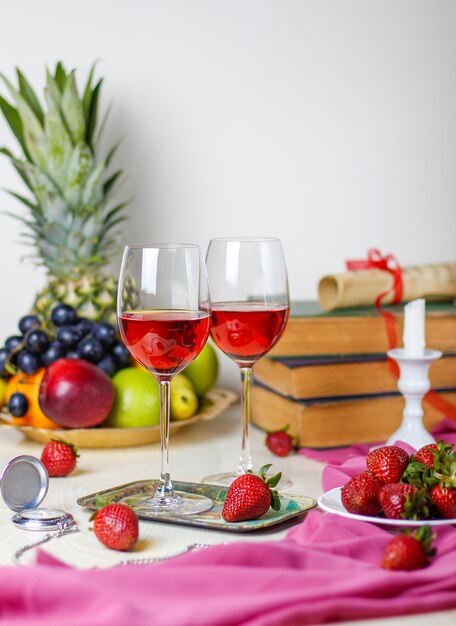 Dos copas de vino rosado en la mesa de madera blanca con libros antiguos y reloj, diferentes frutas tropicales y fresas