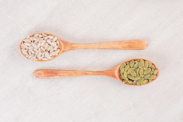 Dos cucharas de madera de semillas de girasol y calabaza sobre superficie blanca.