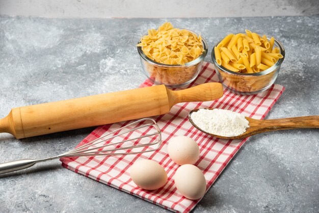 Dos cuencos de pasta cruda, huevos, cuchara de harina y rodillo sobre mesa de mármol con mantel.