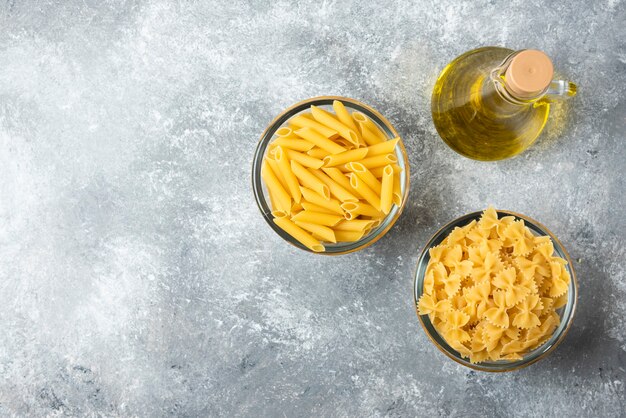 Dos cuencos de pasta penne cruda y farfalle con botella de aceite de oliva sobre fondo de mármol.