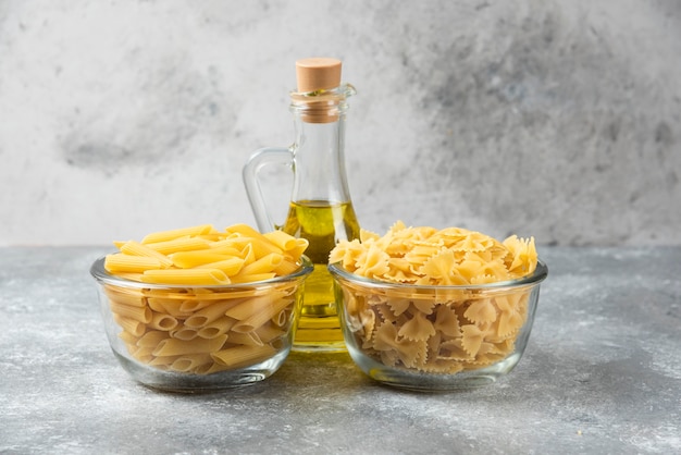 Dos cuencos de pasta penne y farfalle cruda con botella de aceite de oliva en la superficie de mármol.