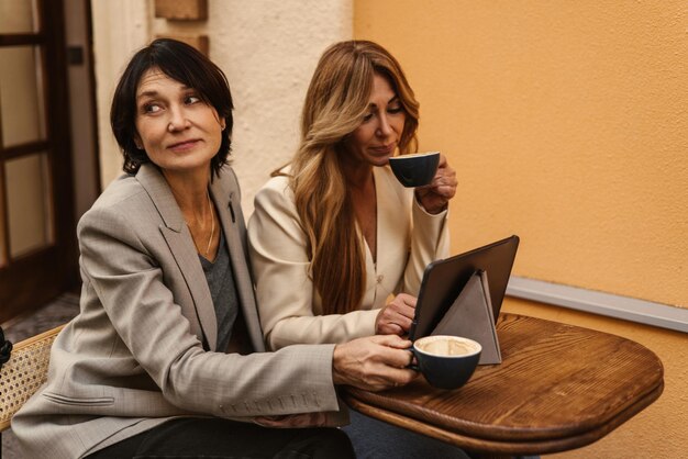 Dos damas caucásicas adultas en una reunión en un café tomando café y usando una tableta en la calle Concepto de tiempo de descanso