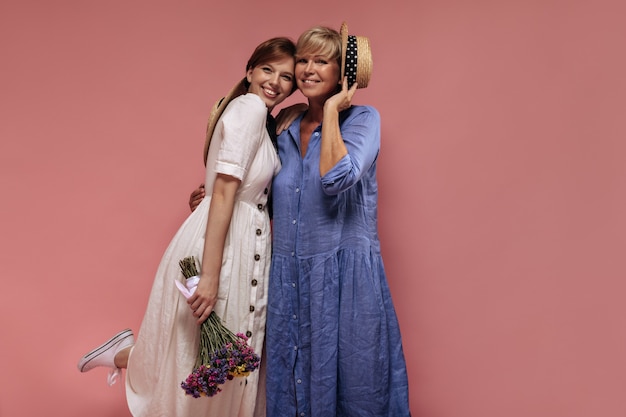 Dos damas modernas con peinado corto fresco en vestidos de moda sonriendo, abrazándose y sosteniendo un ramo y un sombrero de paja sobre un fondo aislado.