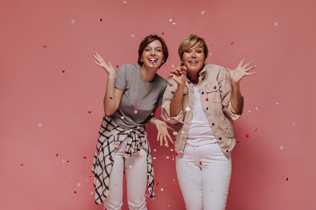 Dos damas positivas con un peinado corto y moderno con un atuendo ligero y elegante sonriendo y posando con confeti sobre fondo rosa