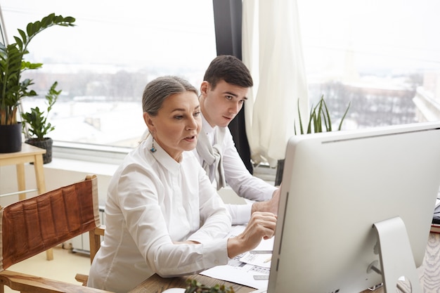 Dos diseñadores caucásicos que trabajan en la oficina moderna usando una computadora genérica: elegante mujer madura compartiendo ideas sobre el diseño de interiores de la sala de estar con su apuesto joven compañero de trabajo. Trabajo en equipo y cooperación