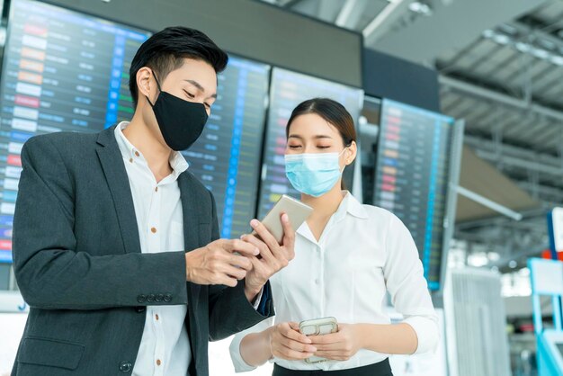 Dos empresarios y empresarias asiáticos con protección de mascarillas en la conversación de la terminal del aeropuerto internacional cerca del concepto de negocio del tablero de información