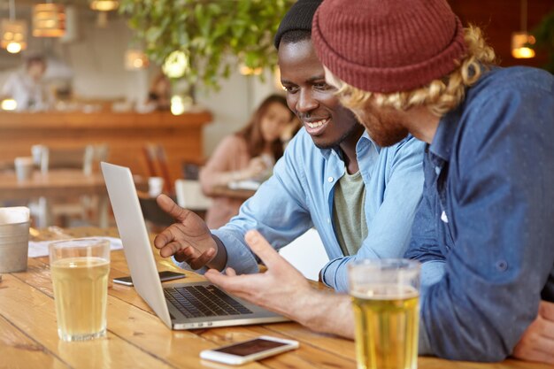 Dos empresarios masculinos con estilo de diferentes razas beben cerveza mientras tienen una reunión de negocios en el bar, discuten un proyecto de inicio común, hablan sobre la estrategia y los planes futuros usando una computadora portátil