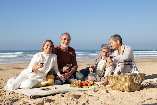 Dos encantadoras parejas mayores disfrutando de un picnic en la playa de arena. Viejos amigos pasando el rato juntos, bebiendo vino y recordando buenos tiempos. Gente sonriendo y mirando a la cámara. Ocio, concepto de amistad