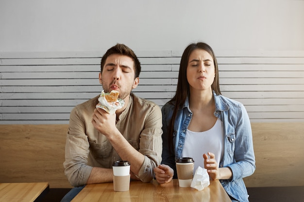 Dos estudiantes hambrientos después de un largo y duro día de estudio cenando en la cafetería. Joven pareja comiendo sándwiches con gran satisfacción.