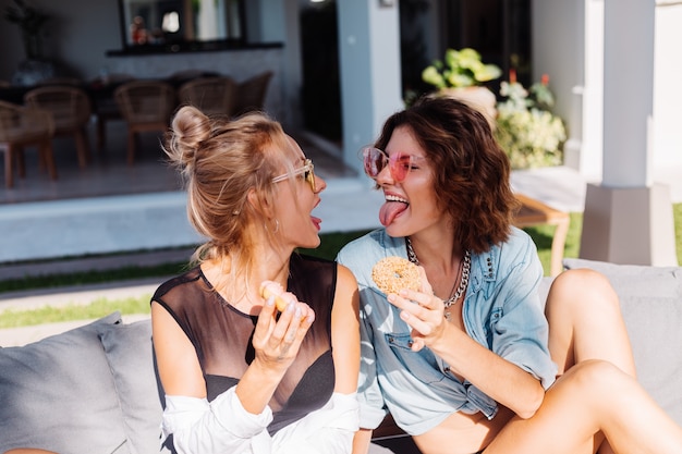 Dos feliz mujer en forma en rosa y amarillo gafas de sol sonriendo divirtiéndose riendo con donas, al aire libre