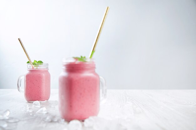 Dos frascos rústicos con batido mezclado fresco de yogur, bayas y hielo aislado sobre mesa de madera y fondo blanco.