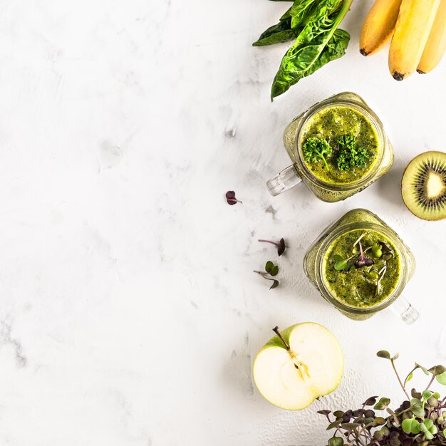 Dos frascos de vidrio de cóctel verde casero a base de espinacas con manzana de plátano y kiwi y verduras húmedas en una vista superior de fondo blanco