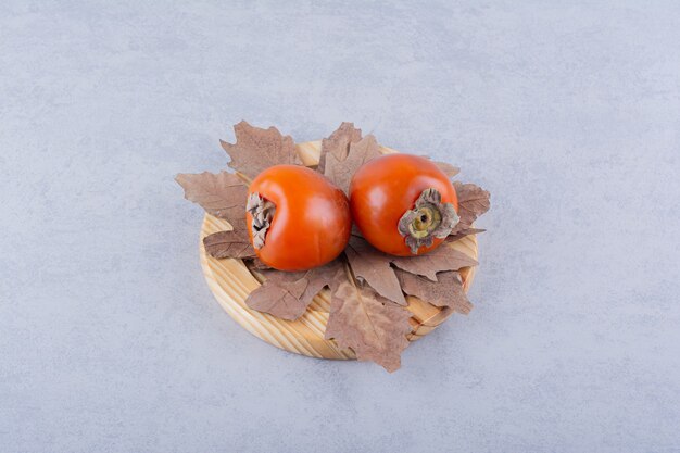 Dos frutas frescas de caqui y hojas secas en placa de madera.