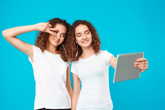 Dos gemelos de mujer haciendo selfie en tableta sobre azul.