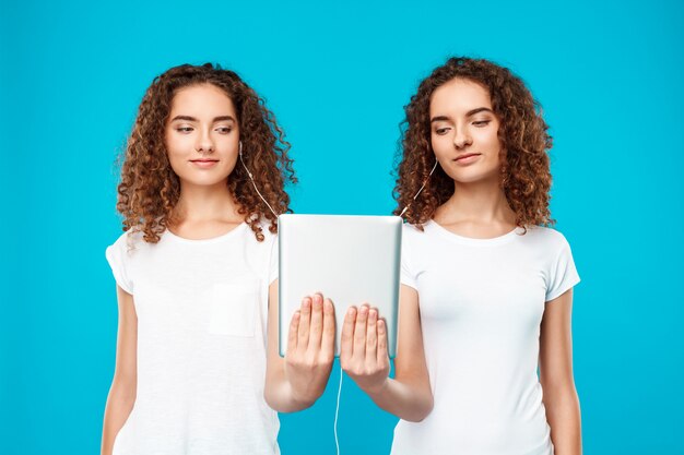 Dos gemelos de mujer mirando tablet, sonriendo sobre azul.