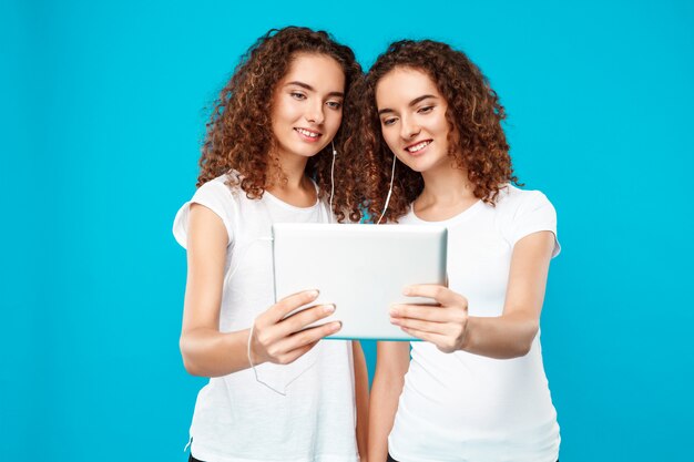 Dos gemelos de mujer mirando tablet, sonriendo sobre azul.