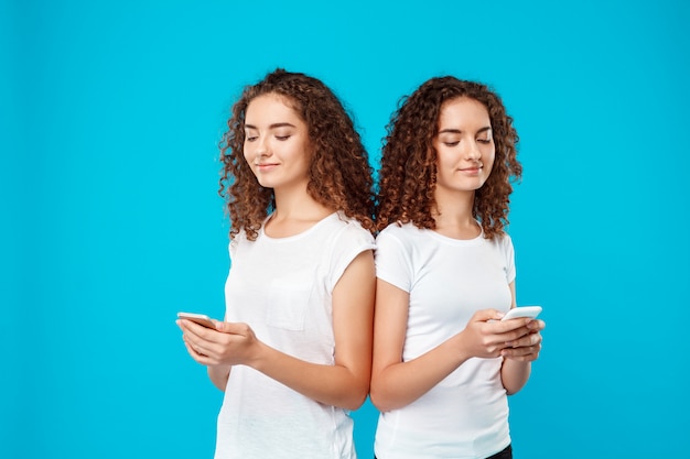 Dos gemelos de mujer sonriendo, mirando los teléfonos sobre azul.