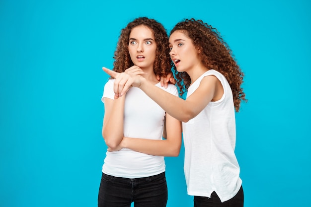 Dos gemelos sorprendidos de la mujer que señalan con el dedo lejos sobre azul.