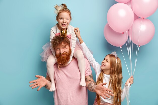 Dos graciosas hermanas traviesas juegan juntas, se hacen cosquillas siendo cuidadas por papá. Niña linda tiene montón de globos de aire. Familia feliz de padre y dos hijas se preparan para las vacaciones, plantean interiores