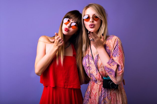 Dos guapas rubias y morenas enviándote besos al aire, espacio de estudio violeta, elegantes vestidos vintage y gafas de sol boho.