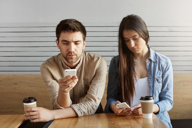 Dos guapos estudiantes se cansaron después del estudio, sentados en la cafetería, tomando café en silencio, mirando a través de las redes sociales en los teléfonos inteligentes.