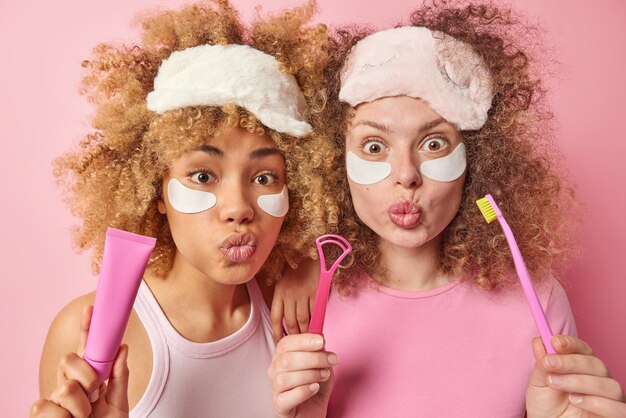 Dos hermanas sorprendidas con el pelo rizado usan máscaras para dormir y aplican parches de belleza debajo de los ojos que van a limpiar los dientes y la lengua con el cepillo parado cerca uno del otro aislado sobre fondo rosa