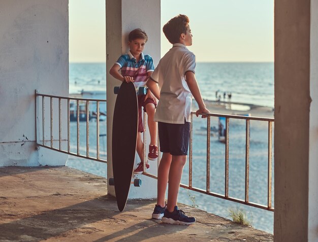 Dos hermanos pequeños parados con una patineta cerca de la barandilla contra el fondo de la costa al atardecer.