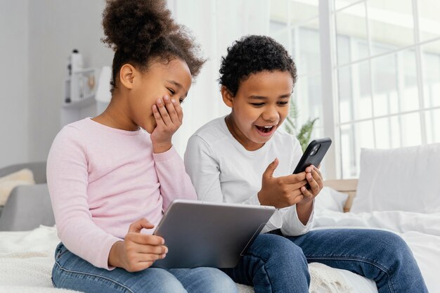 Dos hermanos sonrientes en casa juntos jugando en tableta y teléfono inteligente